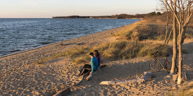 A couple sits on the beach next to wind-blown sea grass as they look out at the water in Kent Island&lt; Maryland, USA