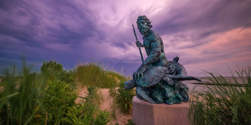 The bronze-turned-blue statue of King Neptune stands grandly among other sealife like turtles and dolphins atop a concrete slab on the sand in Virginia Beach, USA. The sky behind the statue is a mix of purple and pink clouds like paintstrokes in the sky.