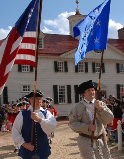 Two paraders holding flags high above their heads and dressed in colonial garb including hats walk down a paved trail in front of George Washington's Mount Vernon in Virginia, USA. Around them are spectators in modern-looking clothes at this reenactment.
